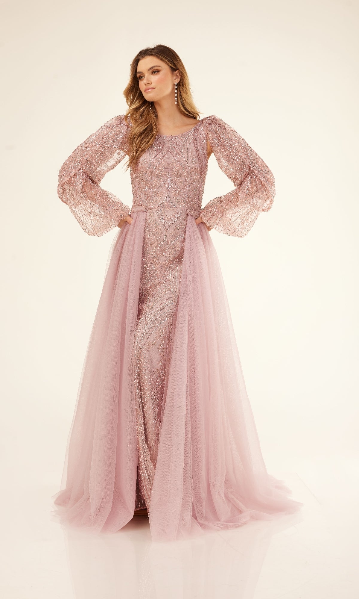 A woman stands against a plain background wearing the Cecilia Couture C273, a long formal gown in light pink featuring sheer embellished sleeves and a flowing tulle skirt.